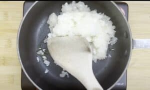 Chopped onions being sautéed in a pan with a wooden spatula on a stovetop for cooking.